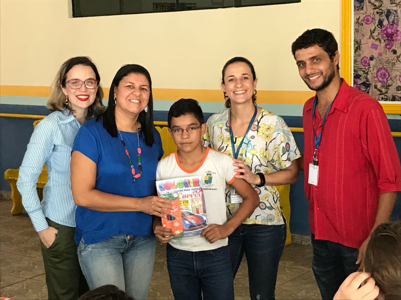 Cinco pessoas, sendo a juíza mais a servidores da Vara e uma criança recebendo exemplar de uma cartilha para colorir.